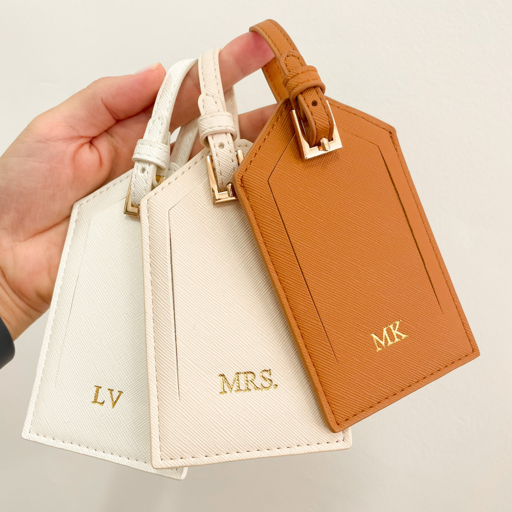 Etiquetas de equipaje personalizadas con iniciales en blanco, camel y beige sostenidas en mano